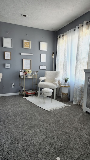 Ein Zimmer mit grauem Teppich, einem weißen, flauschigen Sessel, einem kleinen Beistelltisch mit Pflanze und einer grauen Wand mit gerahmten Motivationssprüchen.