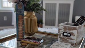 A glass coffee table with a white furry rug, a vase with white flowers, a box of books, and a remote control.