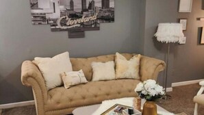 A living room with a beige sofa, a white fluffy ottoman, and a cityscape wall art.