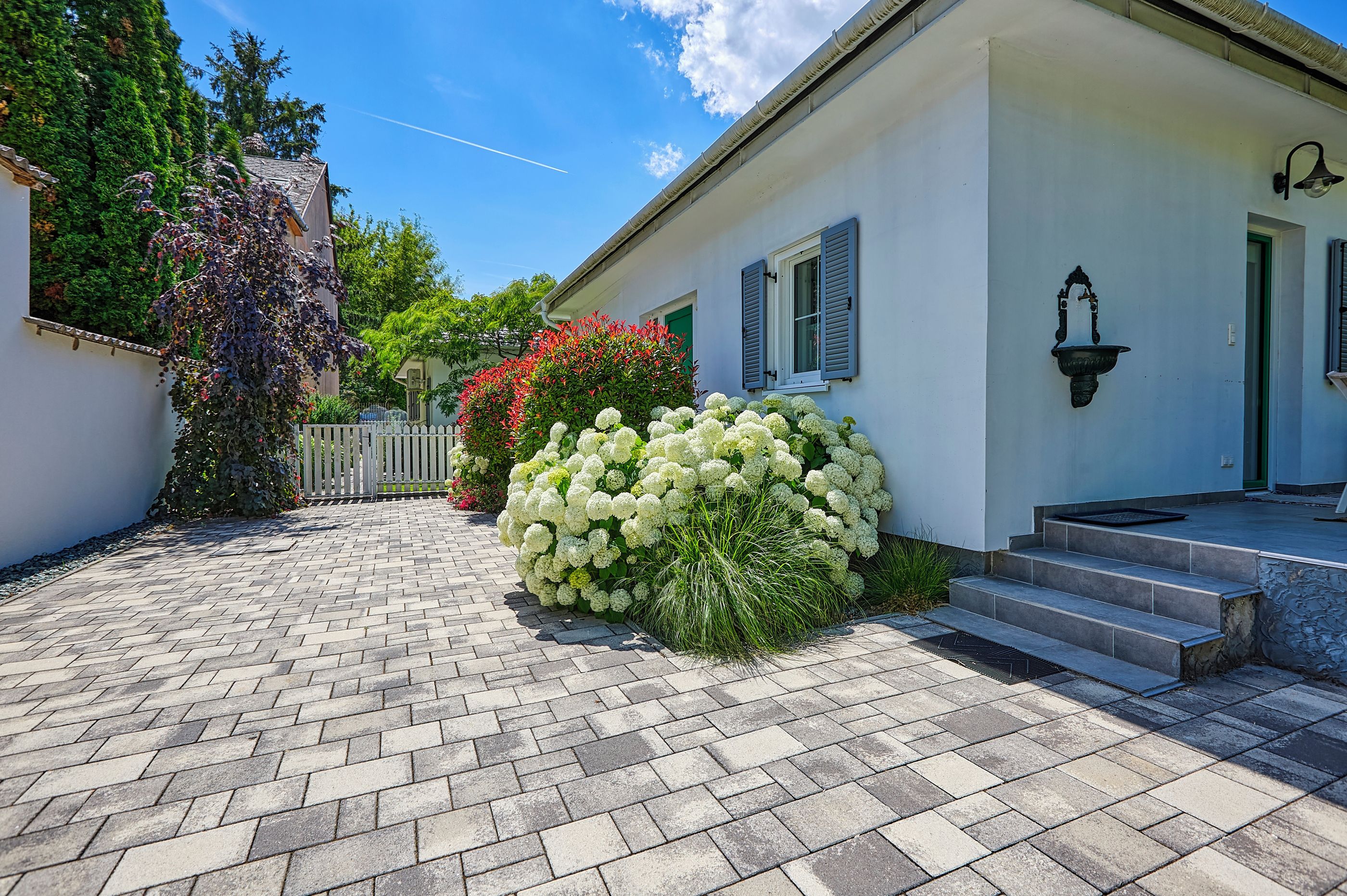 A paved courtyard with a white building, a white picket fence, and lush greenery.