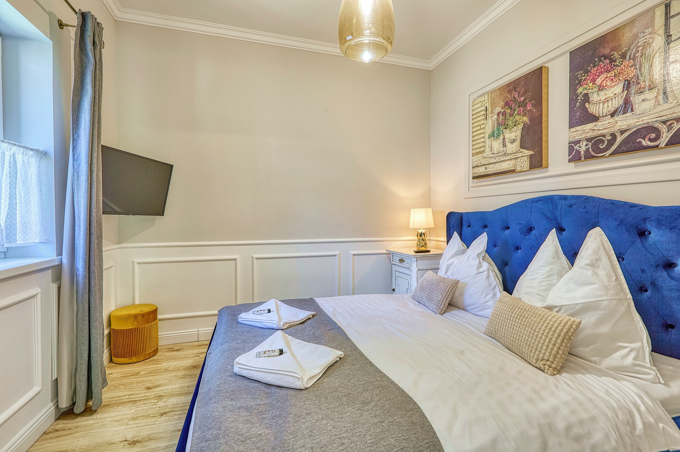 Een hotelkamer met een bed, een nachtkastje met een lamp, een klein krukje en een aan de muur bevestigde televisie.