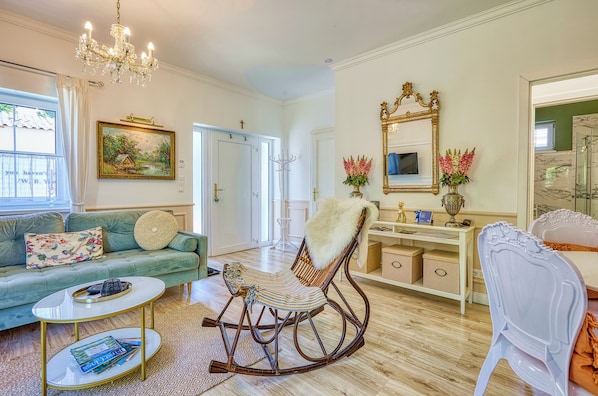 A well-lit living room with a green sofa, a rocking chair, a white chair, a coffee table, and a chandelier.