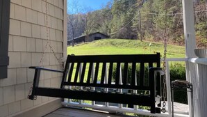 Eine Veranda mit einer schwarzen Bank, einem weißen Geländer und Blick auf ein grünes Feld und Bäume im Hintergrund.