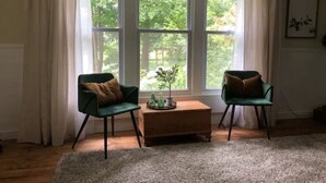 Ein Zimmer mit zwei grünen Stühlen, einem Holztisch und einem großen Fenster mit Vorhängen.