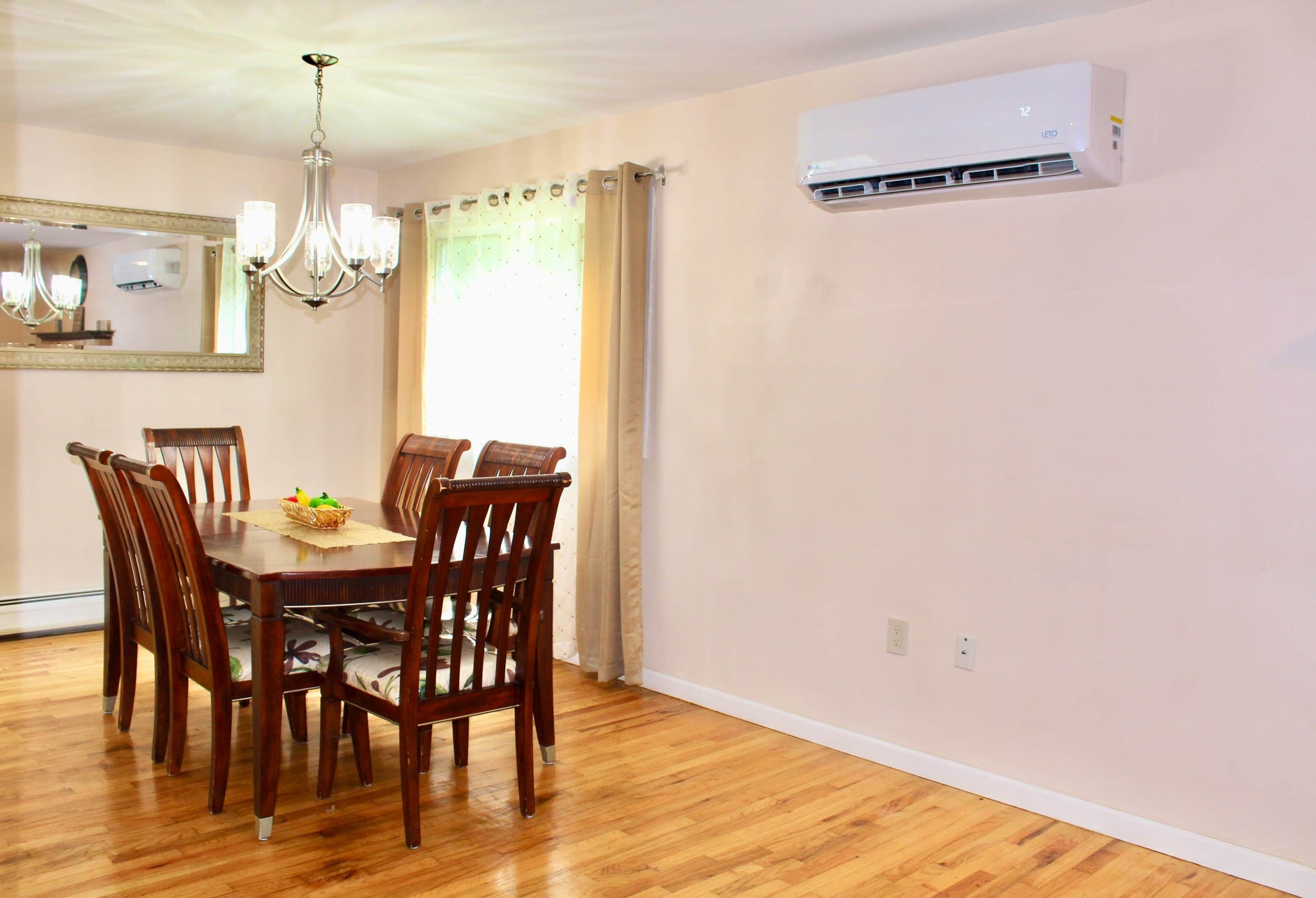 Un comedor con mesa y sillas de madera, un candelabro, un aire acondicionado y una ventana con cortinas.