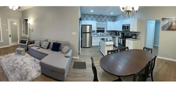 A modern living room with a grey sofa, a dining table, and a kitchen in the background. - All New Deland Den 4BR, pool, blocks fr Stetson & Downtown (DeLand)