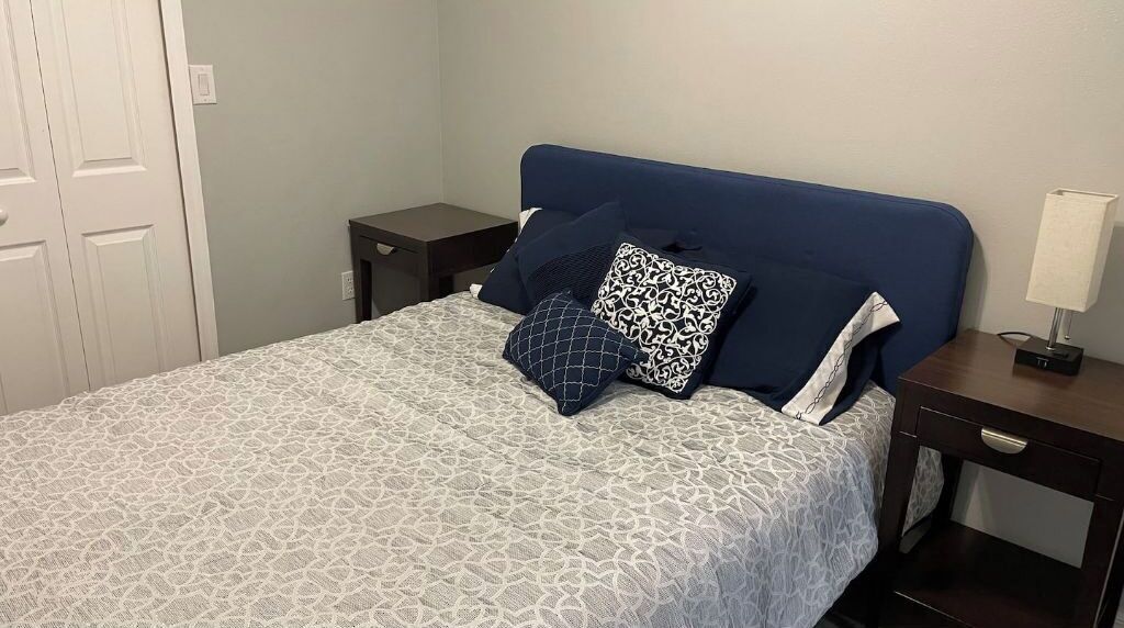 A neatly made bed with a patterned bedspread, a blue headboard, and decorative pillows. There is a bedside table with a lamp and a small drawer.