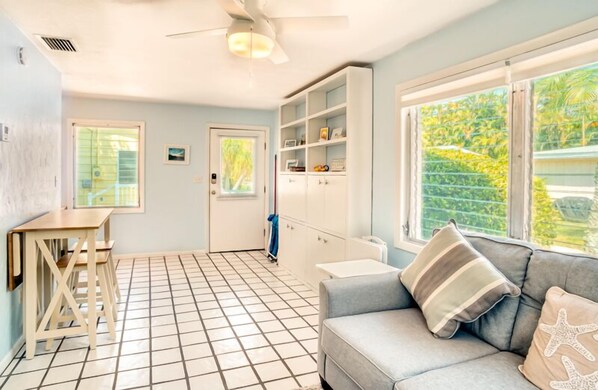 A room with a tiled floor, a sofa with starfish pillows, a ceiling fan, a window with a view of greenery, and a white cabinet with shelves.