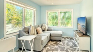 A room with a grey sectional sofa, a small white table, a TV on a wooden stand, and large windows with blinds. - Brand New to the rental market ! (Sanibel)