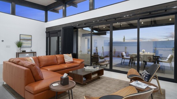 A modern living room with a leather sofa, coffee table, and chairs, large windows with a view of the sea, and a glass door leading to an outdoor seating area.