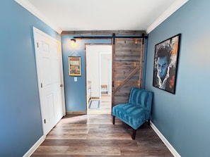 Un couloir avec un mur bleu, un plancher en bois, une chaise bleue, une porte, un luminaire fixé au mur et un tableau encadré.