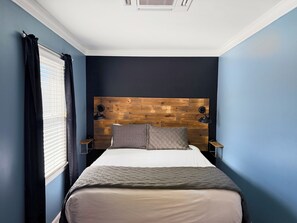 Une chambre à coucher avec une tête de lit en bois, un lit avec un matelas blanc et une literie grise, deux lampes de chevet et une fenêtre avec des stores.
