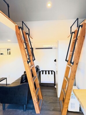 Ein Zimmer mit einer Holzleiter, die an der Wand lehnt, einer schwarzen Ledersofa und einem weißen Klimagerät.