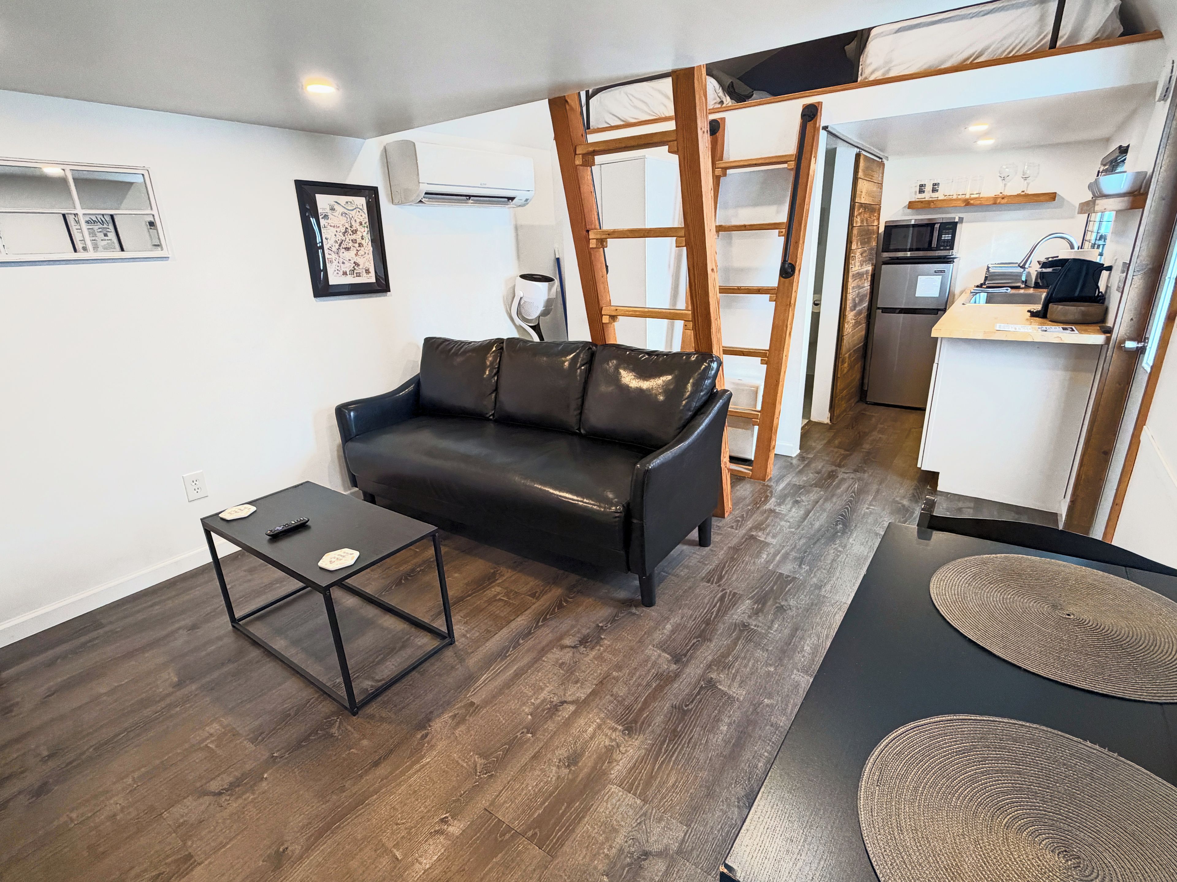 Un espace de vie compact comprenant un canapé en cuir noir, une petite table, une échelle et un coin cuisine.