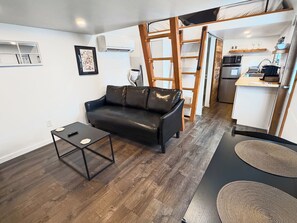 A compact living space with a black leather sofa, a small table, a ladder, and a kitchenette area.