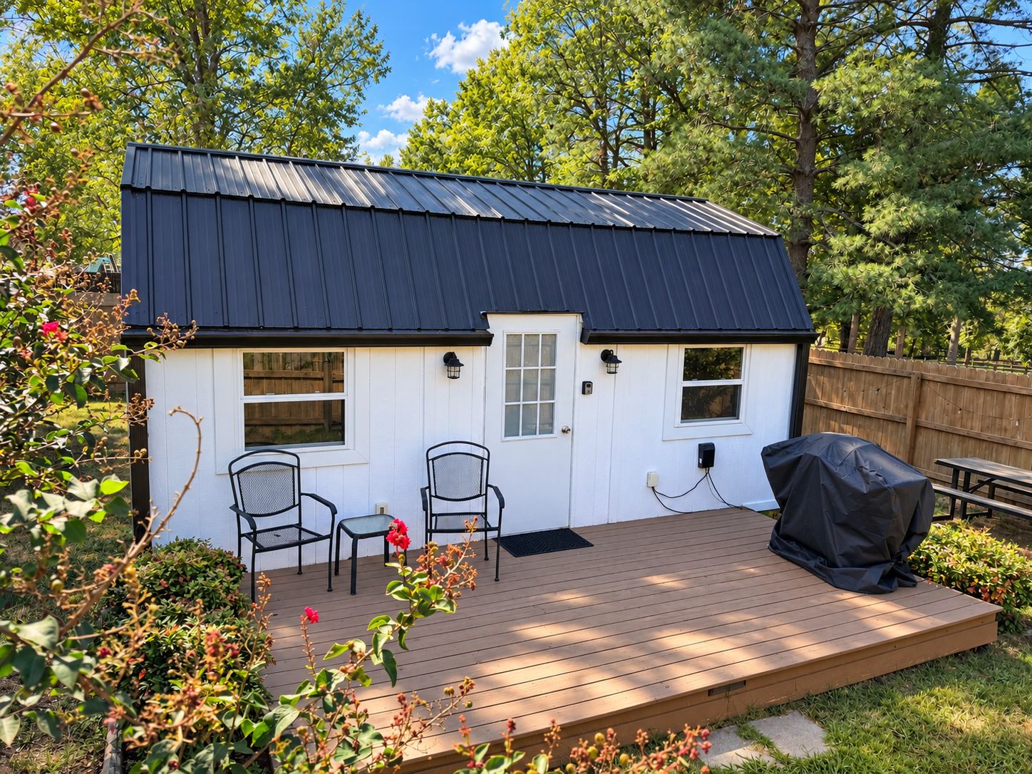 A small white house with a dark roof, two chairs, a table, and a barbecue grill on a wooden deck.