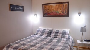 Quarto com colcha xadrez, duas almofadas, quadro emoldurado de uma floresta e abajur.