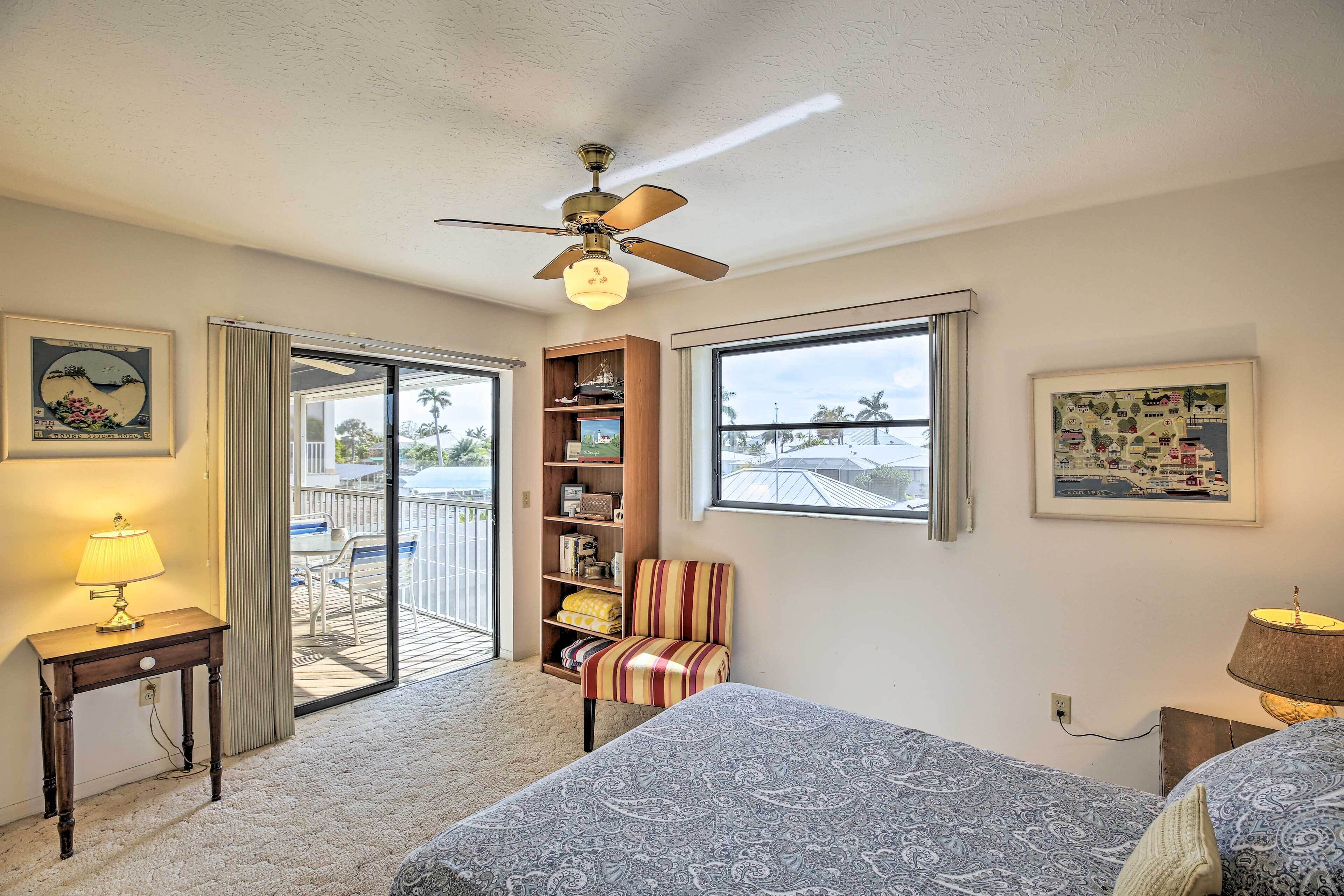 A hotel room with a ceiling fan, a window with a view of palm trees, a bookshelf, a striped armchair, and a bed with a patterned bedspread.