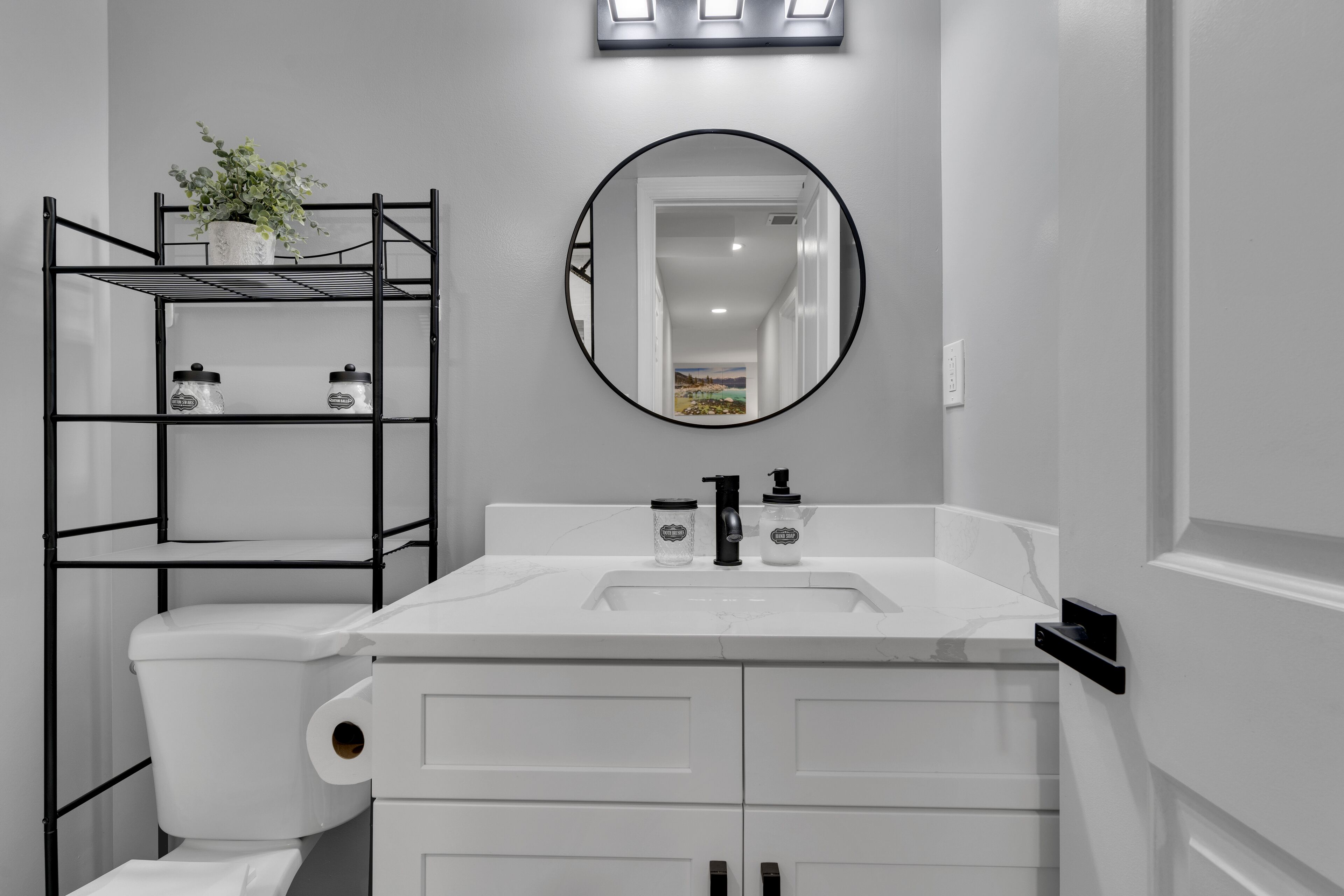 Ein modernes Badezimmer mit einer weißen Toilette, einem weißen Waschbecken mit Spiegel darüber und einer schwarzen Metallregaleinheit mit Vorratsgläsern.