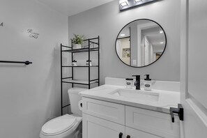 A modern bathroom with a white toilet, a black metal shelf with a potted plant, a white sink with a black faucet, and a round mirror. - Cozy Modern Beautiful Studio+Parking. Walk to DC Metro. Perfect for Long Stays! (Washington)