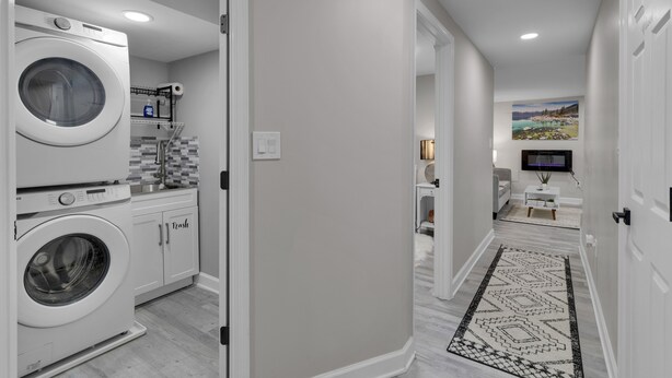 A laundry room with stacked washers and dryers, a tiled backsplash, and a trash bin. - Cozy Modern Beautiful Studio+Parking. Walk to DC Metro. Perfect for Long Stays! (Washington)