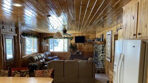 L’intérieur chaleureux d’un chalet avec un plafond en bois, une cheminée en pierre, un grand canapé et un coin cuisine.