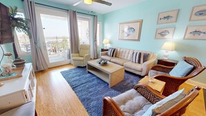 A living room with a sofa, armchairs, a coffee table, and a dining table. There are framed fish pictures on the wall and a ceiling fan. - Ocean Views & Central location~ Pier Point Villas B202 (Folly Beach)