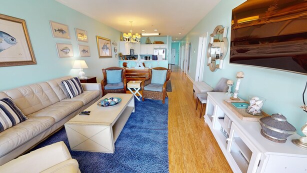 A living room with a sofa, coffee table, and chairs, a kitchen area with a dining table, and a hallway leading to a bedroom. - Ocean Views & Central location~ Pier Point Villas B202 (Folly Beach)