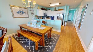 A dining area with a wooden table and benches, a chandelier, and a kitchen in the background. - Ocean Views & Central location~ Pier Point Villas B202 (Folly Beach)
