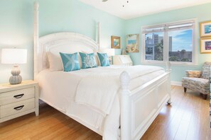 A bedroom with a white four-poster bed, a nightstand with a lamp, a chair, and a window with a view of buildings. - Ocean Views & Central location~ Pier Point Villas B202 (Folly Beach)