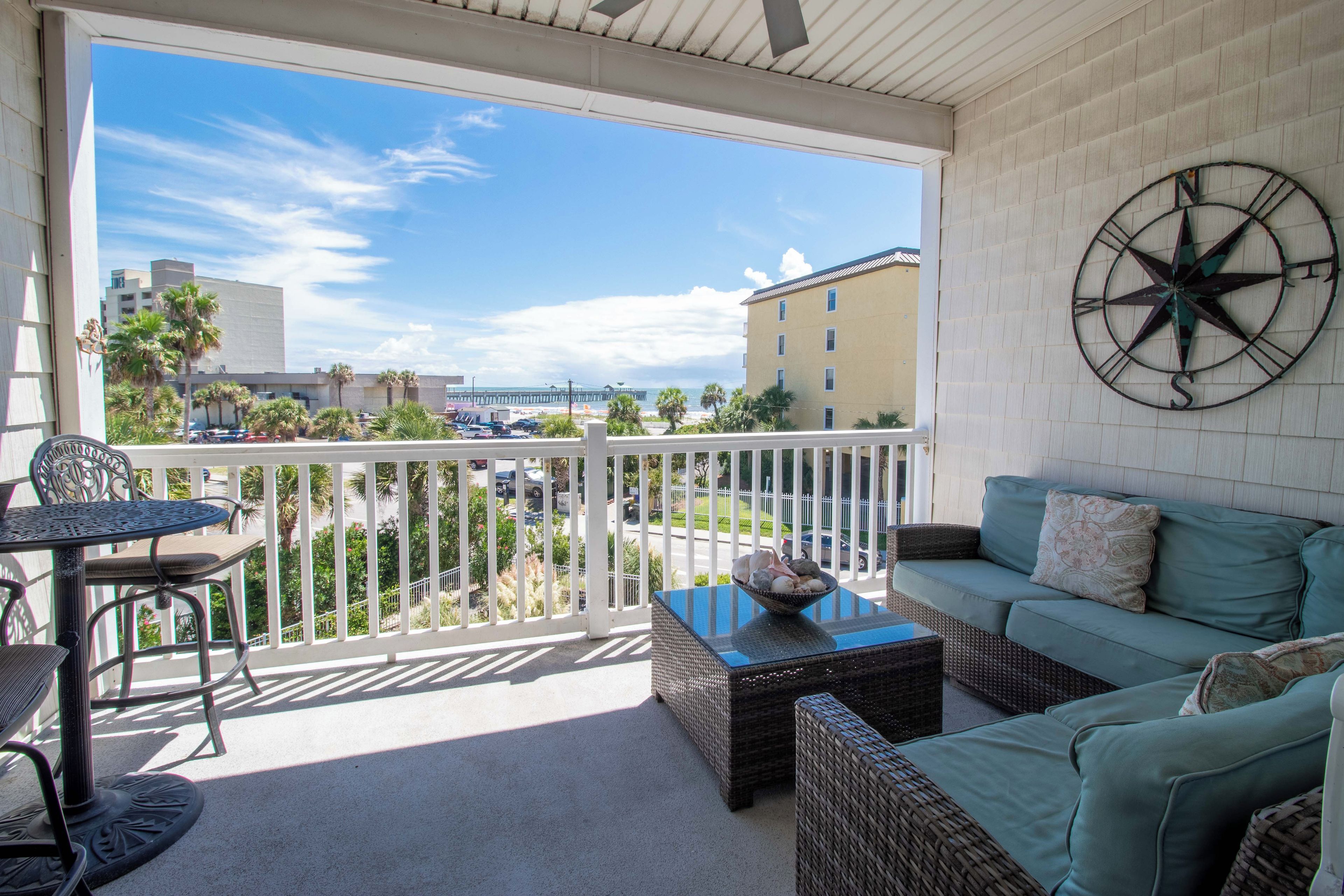 A balcony with a view of the beach and cityscape, furnished with a sofa, table, and chairs.