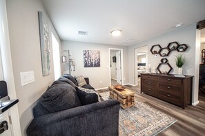 Un salon avec un canapé gris, une table basse en bois et un tapis à motifs. On y trouve une commode en bois foncé et un mur orné de décorations hexagonales.