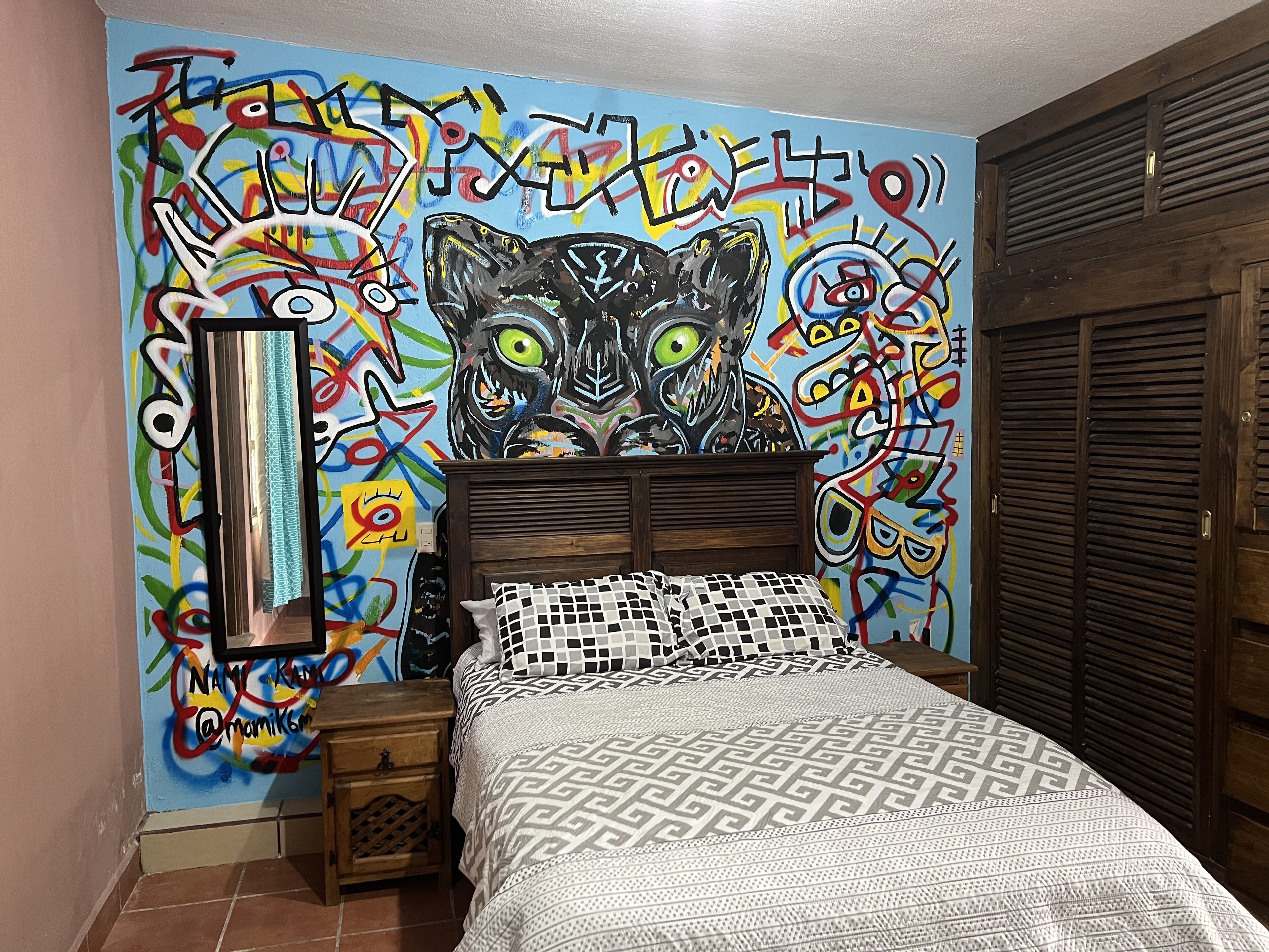 Ein Schlafzimmer mit einem farbenfrohen Graffiti-Wandbild, einem Holzkopfteil, einem Bett mit gemusterten Betttextilien und einem Nachttisch mit Spiegel.