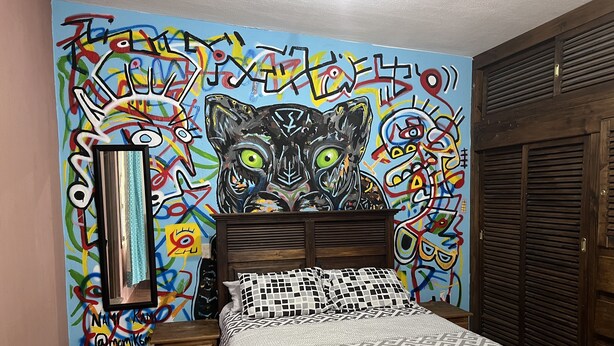 Een slaapkamer met een levendige graffiti-muurschildering, een houten hoofdbord, een bed met beddengoed met een patroon en een nachtkastje met een spiegel.