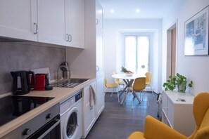Uma cozinha moderna com máquina de lavar, cafeteira e chaleira vermelha. Há uma área de refeições com mesa branca e cadeiras amarelas.