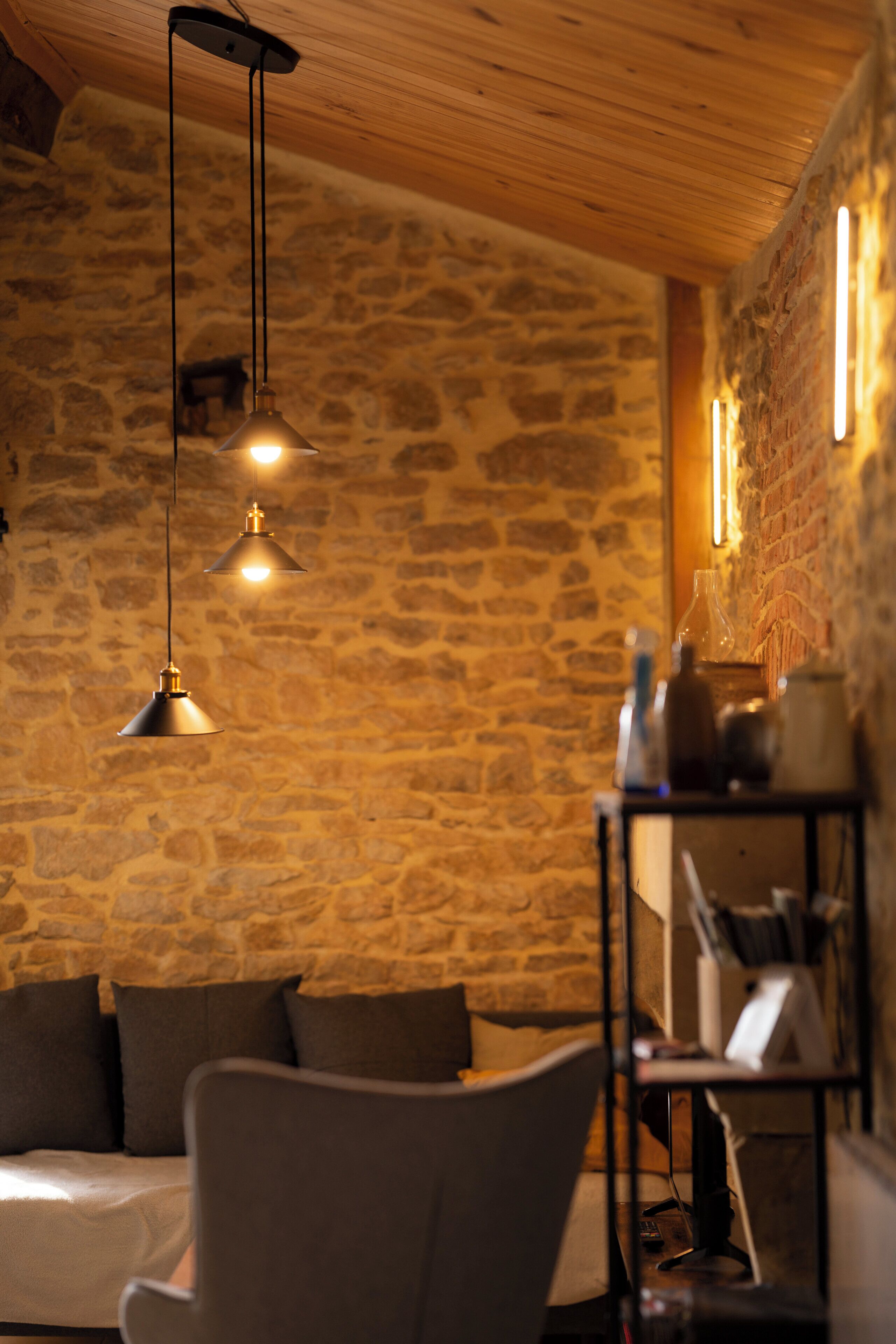 Um quarto aconchegante com parede de pedra, teto de madeira e um sofá com almofadas. Há luminárias pendentes no teto.