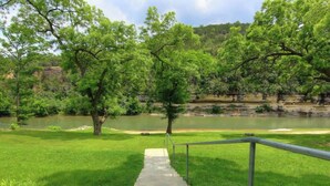 A concrete staircase leading to a grassy area with a metal railing, surrounded by trees and a body of water. - RG 306 Hillside River Retreat (New Braunfels)