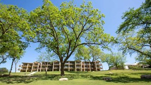 A multi-story building with a flat roof, surrounded by a grassy area with trees. - RG 306 Hillside River Retreat (New Braunfels)