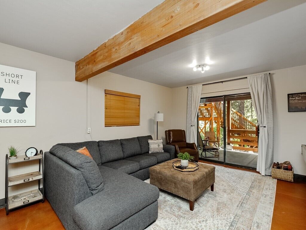 Una sala de estar con un sofá seccional gris, un reposapiés marrón, una viga de techo de madera, una ventana con cortinas y un letrero en la pared.