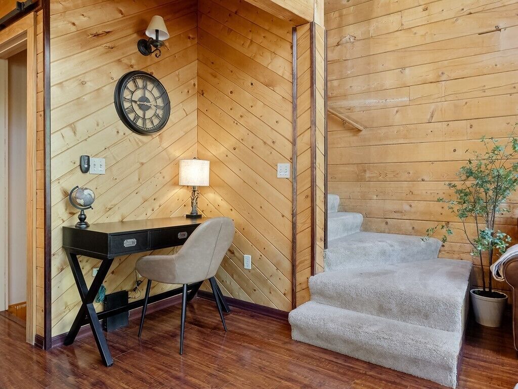 A cozy room with wooden walls, a desk with a lamp, a globe, and a chair. There are stairs leading to an upper level.