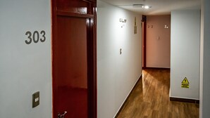 Un pasillo con piso de madera, una puerta roja con la etiqueta '303' y un letrero de precaución amarillo en la pared.