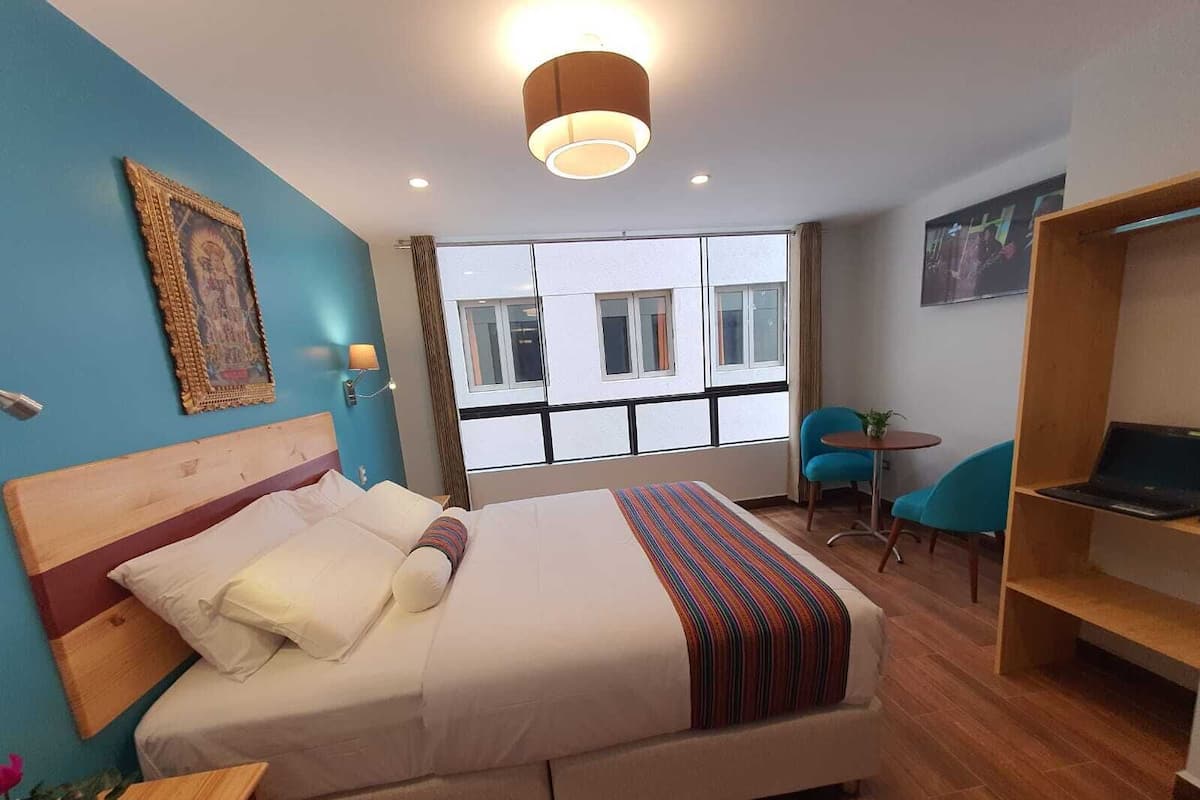 A hotel room with a bed, a desk, a chair, a small table, and a window with a view of buildings.