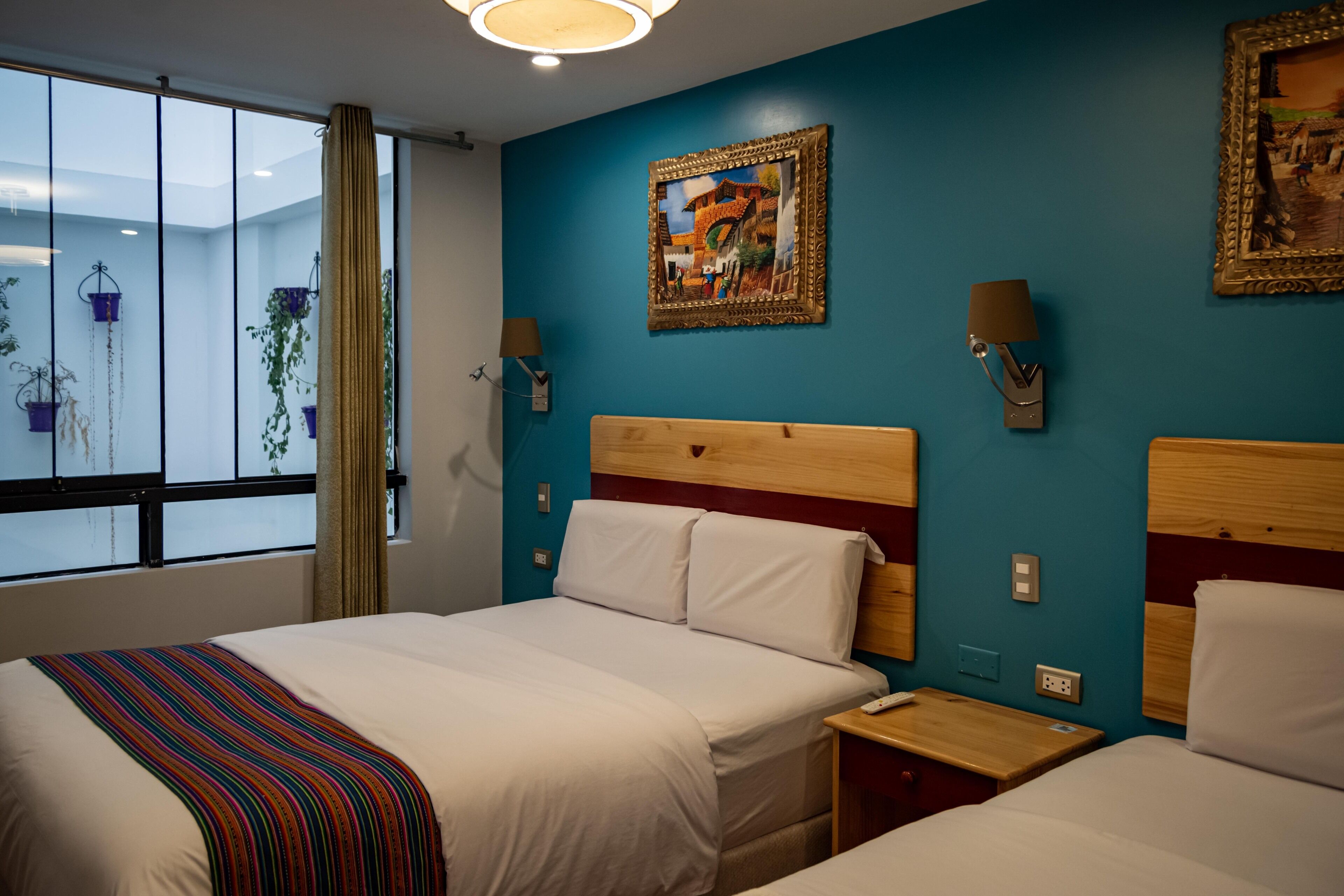 A hotel room with two beds, a large window with curtains, a painting on the wall, and a bedside table with a lamp.