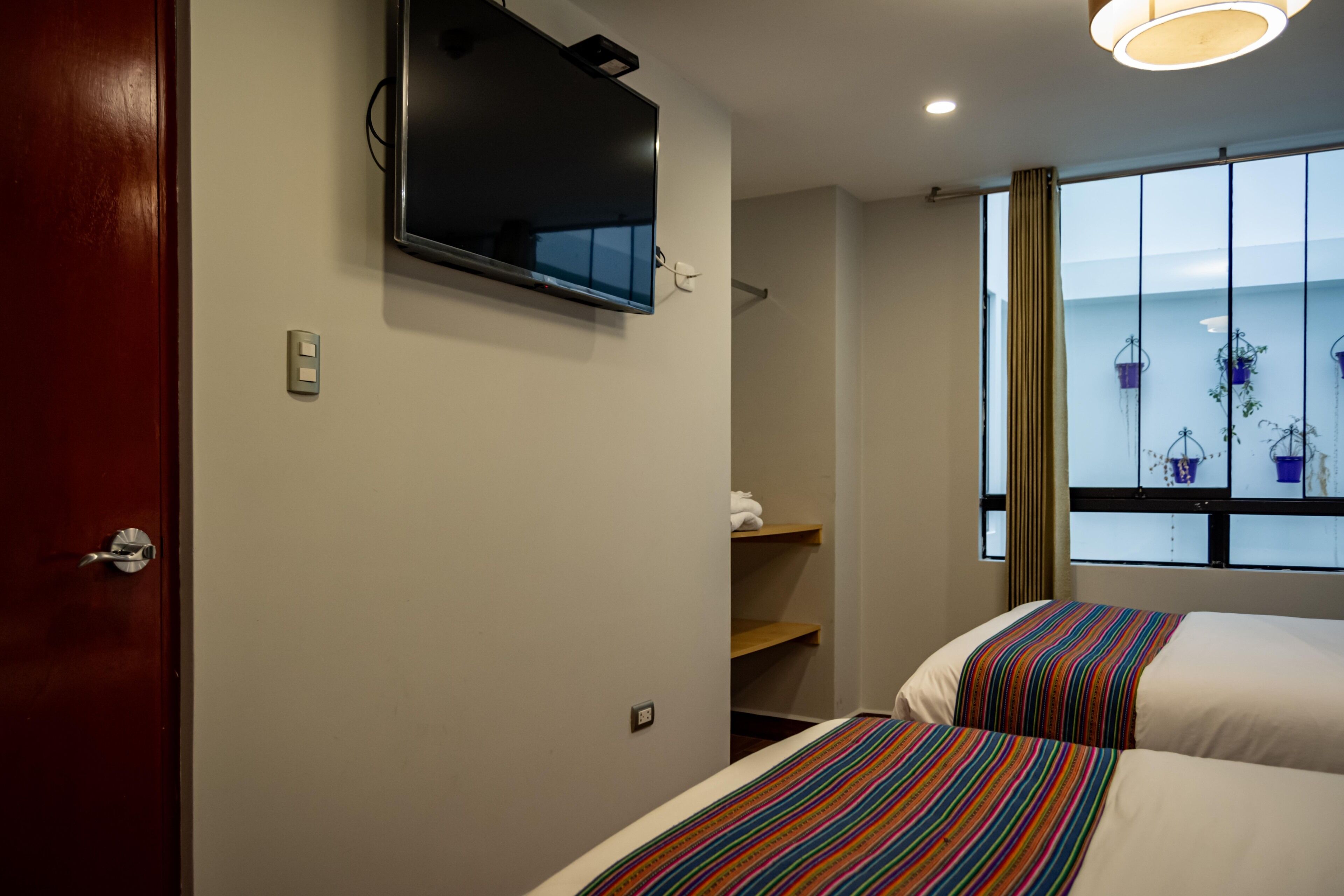 A hotel room with a TV mounted on the wall, a bed with striped bedding, a window with curtains, and a shelf with towels.