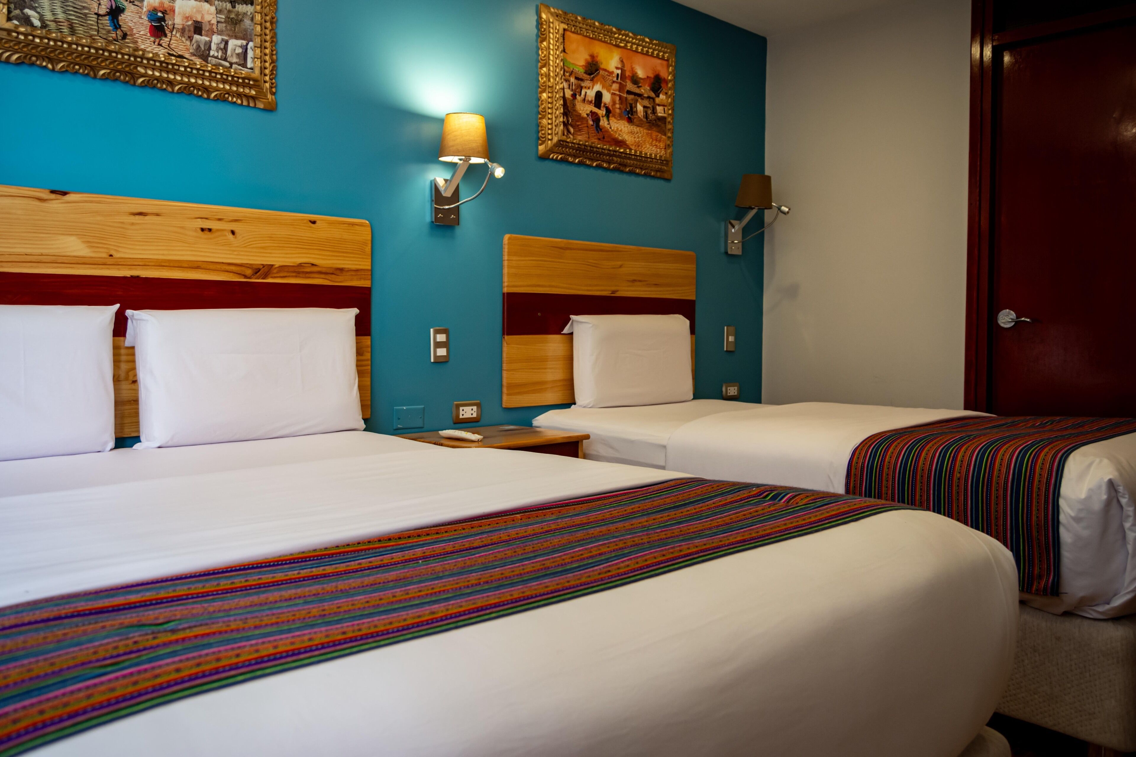 Une chambre d’hôtel avec deux lits, une tête de lit en bois, une couvre-lit rayée aux couleurs vives, une lampe fixée au mur et une porte.