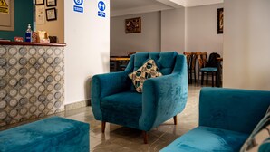 Un moderno salón con un sillón azul y reposapiés, una pared con estampado, una zona de comedor al fondo y un mostrador de recepción con un letrero azul.