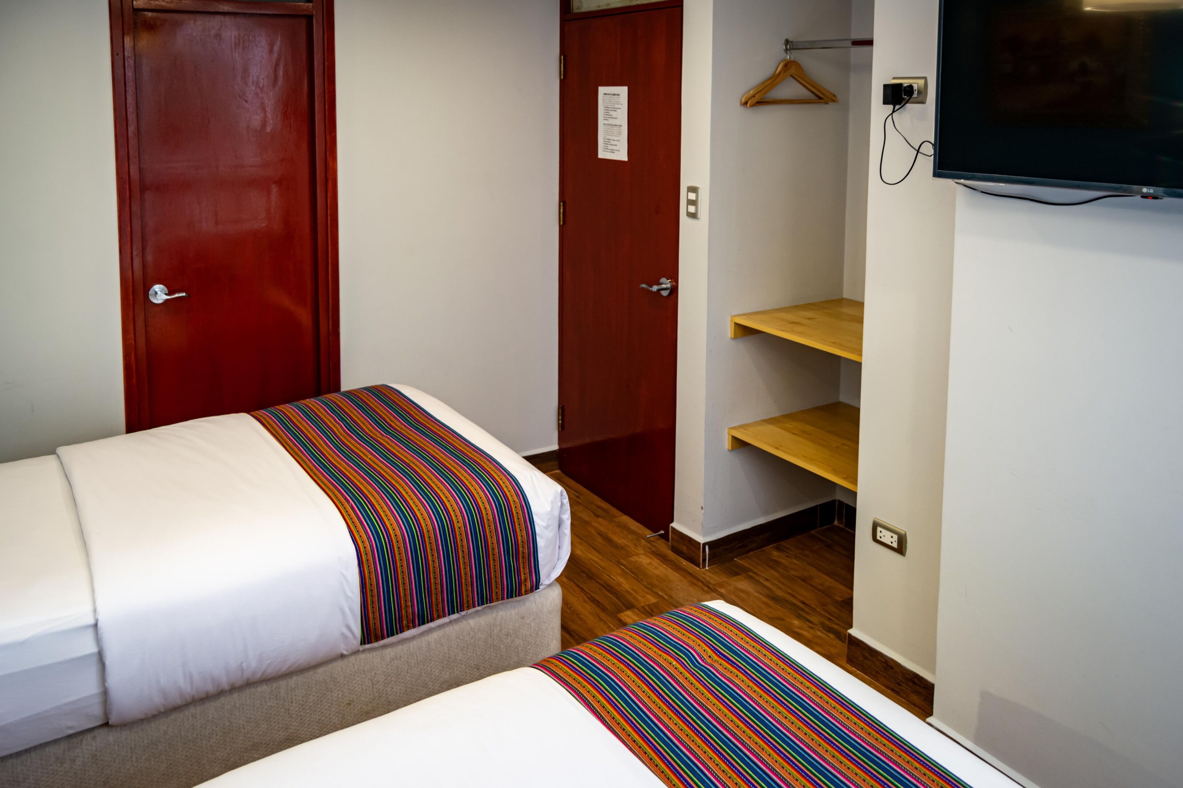 A hotel room with two beds, a TV mounted on the wall, a wooden shelf, and a red door.
