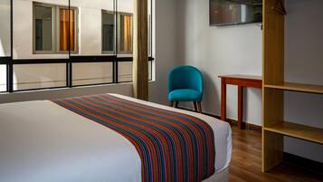 A hotel room with a large bed, a blue chair, a wooden shelf, and a television on the wall.
