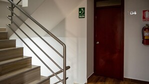 Una escalera con barandilla de metal, una puerta roja, un letrero de salida de emergencia verde y un extintor de incendios fijado en la pared.