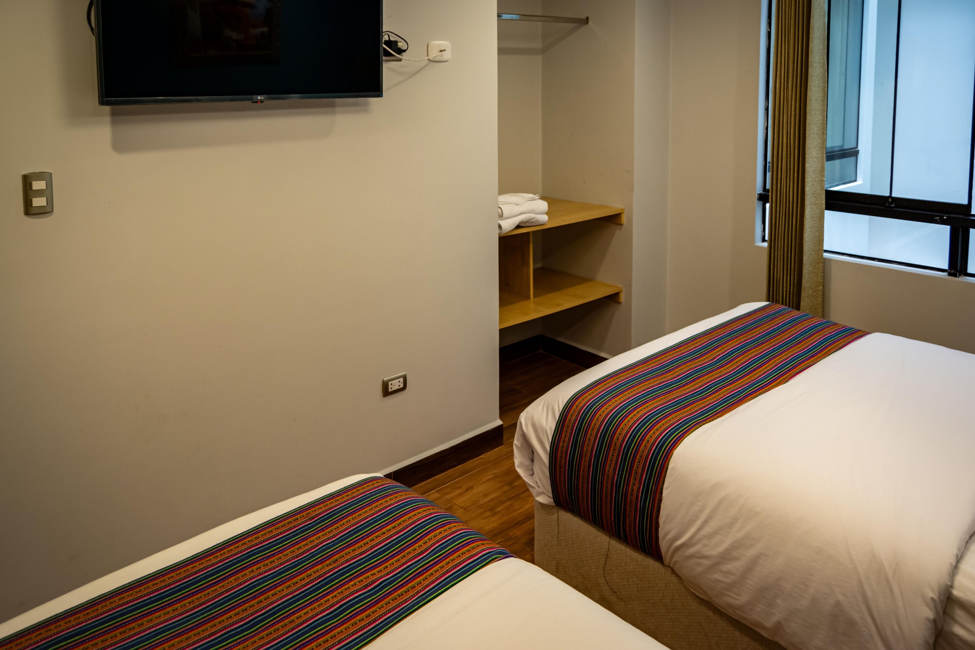 A hotel room with two beds, a television mounted on the wall, and a window with curtains.